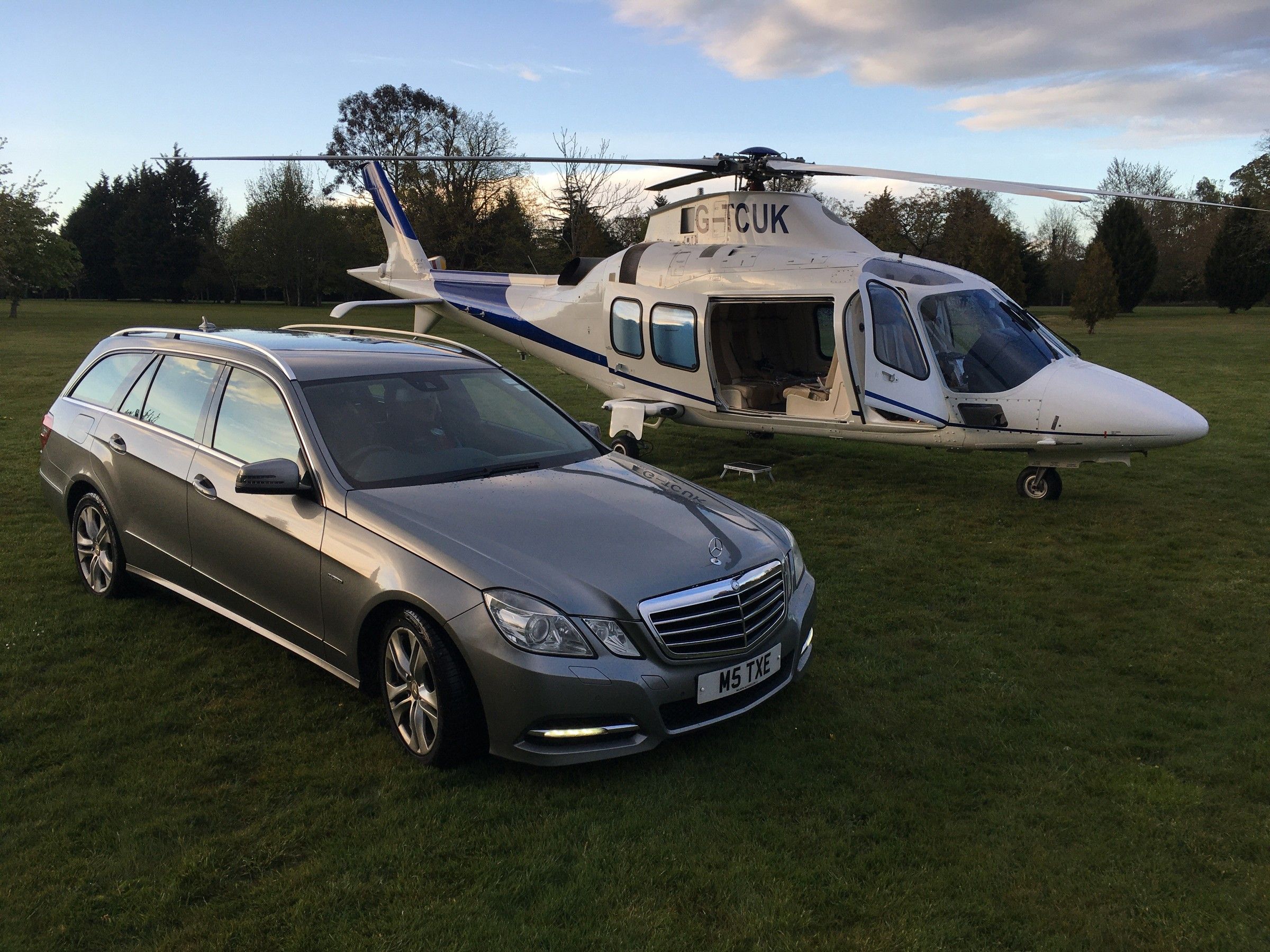 Silver Mercedes-Benz E-Class estate on grass beside a helicopter